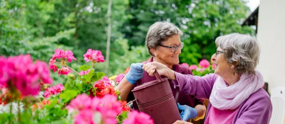 Seniorin mit Pflegefachkraft im Garten beim Blumengießen